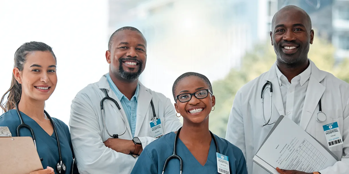 2 women and 2 men in doctor or nurse uniforms smiling at the camera, some holding charges or clip boards, wearing stethoscopes around their necks