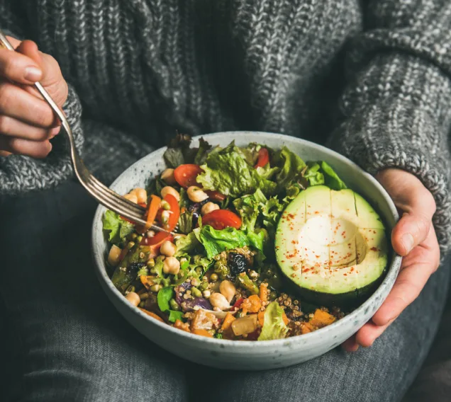 Healthy vegetarian dinner. Woman in jeans and warm sweater holding bowl with fresh salad, avocado, grains, beans, roasted vegetables, close-up. Superfood, clean eating, vegan, dieting food concept