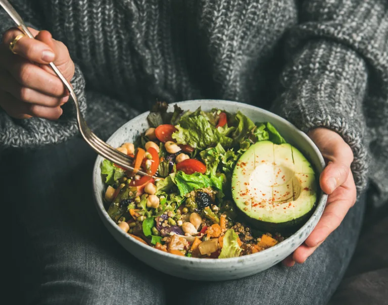 Healthy vegetarian dinner. Woman in jeans and warm sweater holding bowl with fresh salad, avocado, grains, beans, roasted vegetables, close-up. Superfood, clean eating, vegan, dieting food concept